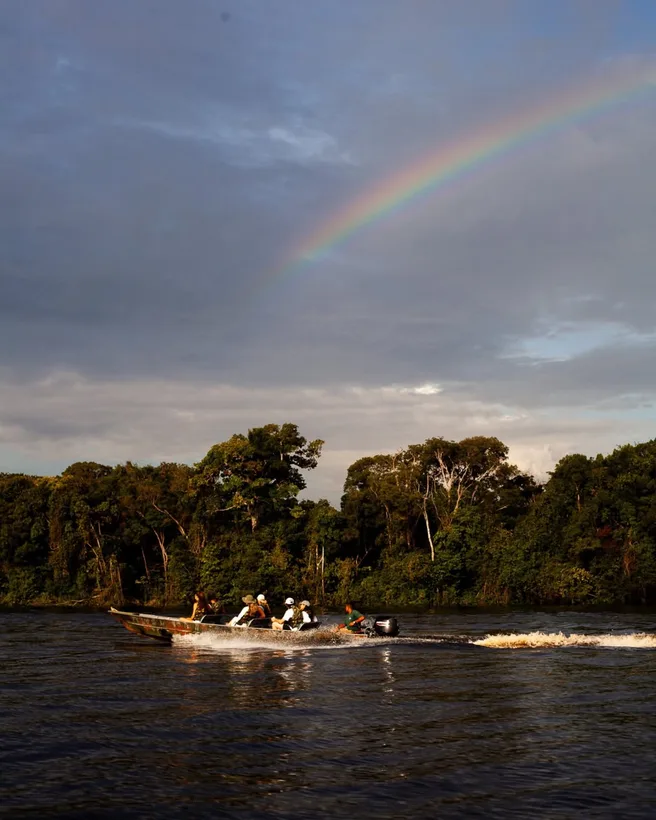 Canoa com pessoas no rio ao anoitecer, com um arco-íris no céu nublado