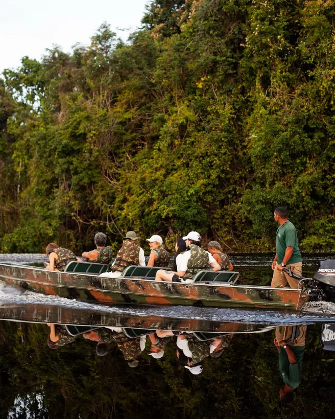 Canoa comprida cheia de gente refletida na água parada, com a mata atrás